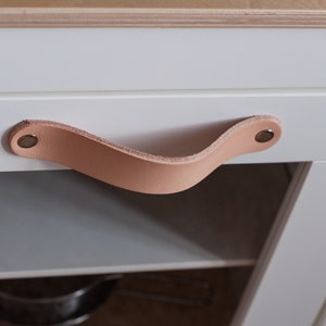 May include: A light brown leather cabinet pull with brass rivets. The pull is curved and has a simple design.