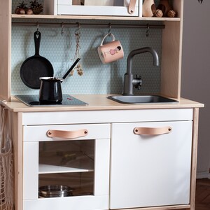 May include: A white wooden play kitchen with a black stovetop, a sink, and two cabinets with leather handles. The kitchen has a white microwave with a glass door and a shelf above the sink. The kitchen is on legs and has a light blue and white patterned backsplash.