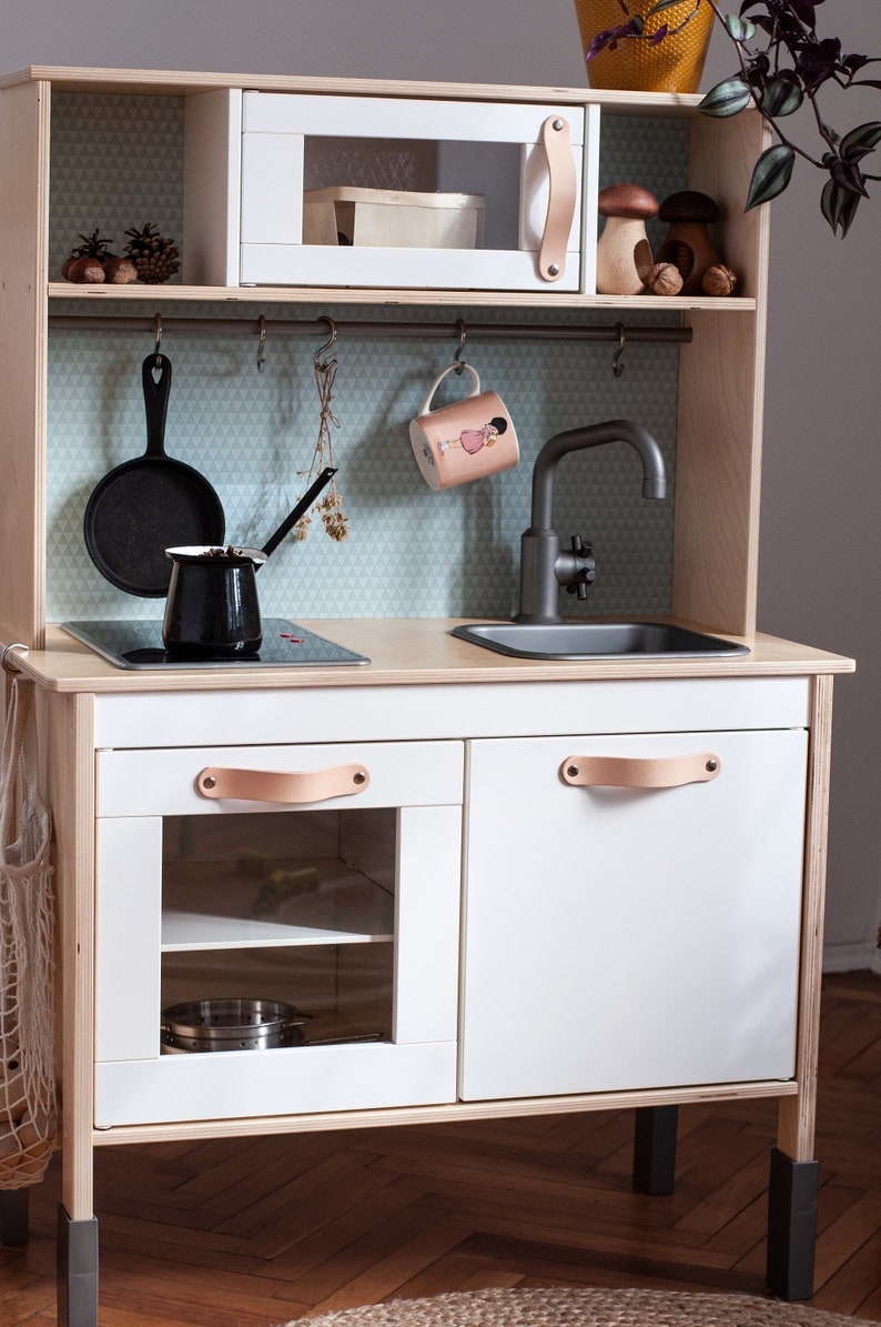 May include: A white wooden play kitchen with a black stovetop, a sink, and two cabinets with leather handles. The kitchen has a white microwave with a glass door and a shelf above the sink. The kitchen is on legs and has a light blue and white patterned backsplash.