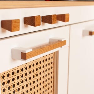 以下が含まれることがあります： 木製のノブと籐の織り込まれたパネルが付いた白いキッチンのキャビネット。キャビネットのドアには、木製のアクセントが付いた白いハンドルが付いています。