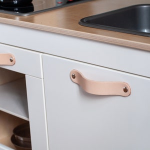 May include: White play kitchen with a sink, stovetop, and two cabinets. The cabinets have leather handles with metal rivets.