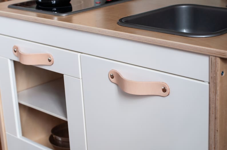 May include: White play kitchen with a sink, stovetop, and two cabinets. The cabinets have leather handles with metal rivets.