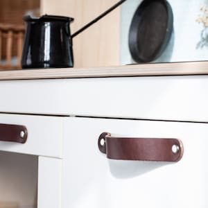 May include: White kitchen cabinets with brown leather handles. The cabinets have a modern design and are perfect for a play kitchen.