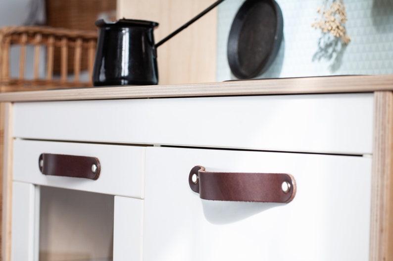 May include: White kitchen cabinets with brown leather handles. The cabinets have a modern design and are perfect for a play kitchen.