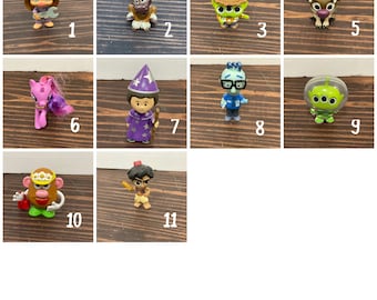 May include: A collection of 11 small plastic figurines, including a purple unicorn, a green alien, a brown reindeer, a blue character with glasses, a brown character with a turban, a brown character with a hat, a purple character with a hat, a brown character with antlers, a pink character with a bow, a brown character with a wrench, and a green character with a hat.