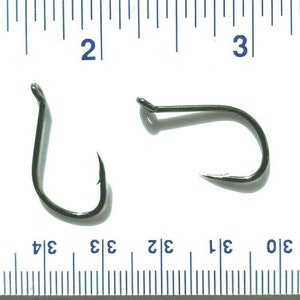 Puede incluir: Dos anzuelos de pesca negros con una regla en el fondo. Los anzuelos miden aproximadamente 2,5 cm de largo.