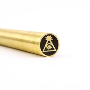Pode incluir: Uma haste de metal dourada com um design circular preto e dourado na extremidade. O design apresenta um olho dentro de um triângulo com uma estrela em forma de explosão acima.