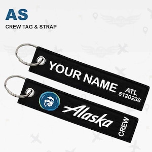 Op de afbeelding: Twee zwarte crew tag en strap sleutelhangers. Eén sleutelhanger leest "YOUR NAME" en "ATL 5120236". De andere sleutelhanger heeft het Alaska Airlines logo en de woorden "Alaska" en "CREW". Beide hebben zilveren sleutelringen.