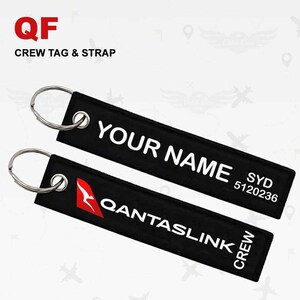 Op de afbeelding: Twee zwarte Qantaslink crew tag & strap sleutelhangers met zilveren ringen. Eén tag leest "YOUR NAME" en "SYD 5120236". De andere tag heeft het Qantas logo en leest "QANTASLINK CREW". De linkerbovenhoek leest "QF CREW TAG & STRAP".