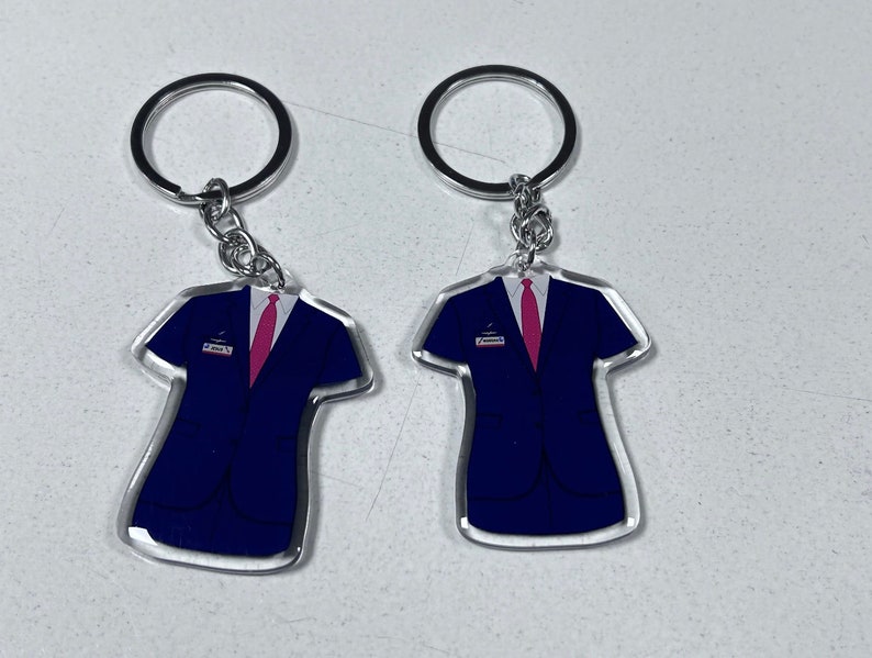 May include: Two clear acrylic keychains shaped like a suit jacket with a pink tie. The keychains are attached to silver key rings.