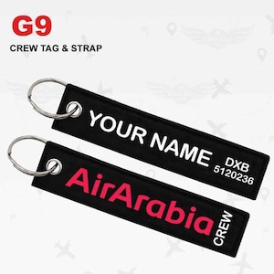 May include: Two black crew tag and strap keychains with silver key rings. One keychain has the text "YOUR NAME" in white. The other keychain has the text "AirArabia" in red. The top left corner has the text "G9 CREW TAG & STRAP".