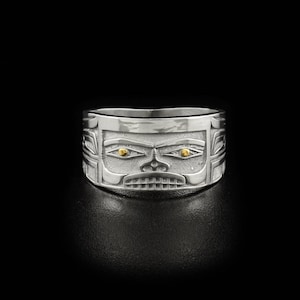 May include: A silver ring with a detailed face design and gold eyes. The ring is wide and has a textured surface.