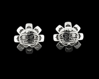 May include: A pair of silver-toned stud earrings. Each earring features a stylized sun face design with a central face surrounded by petal-like shapes. The earrings have a detailed, engraved appearance and are set against a black background.