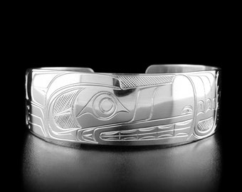 May include: A silver bracelet with a detailed carving of a stylized animal face. The design is reminiscent of Northwest Coast Indigenous art.