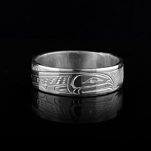 May include: A polished silver band ring featuring an intricate engraved design of a stylized bird with curved lines and cross-hatched textures. The metal surface is reflective, showing a clear mirror image of the ring on the dark surface below.