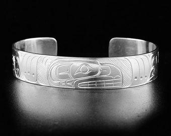 May include: A silver cuff bracelet with a detailed Native American design. The bracelet features a central engraved image of a stylized bird or animal, surrounded by intricate patterns. The open-ended design allows for easy wear.