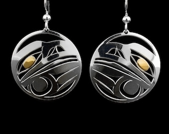 May include: A pair of silver earrings with a circular design featuring a stylized raven with gold accents. The earrings are made of sterling silver and are approximately 1.5 inches in diameter.