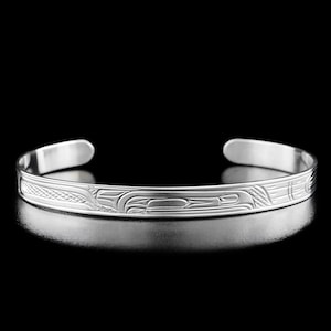 May include: A silver bracelet featuring an intricate design of a bird, possibly a raven, in a Northwest Coast style. The bracelet is crafted from polished metal and has a smooth, rounded surface.