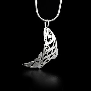 Op de afbeelding: Een zilveren hanger in de vorm van een gestileerde zalm, met ingewikkelde details en een uitgesneden ontwerp. De hanger hangt aan een zilveren ketting.