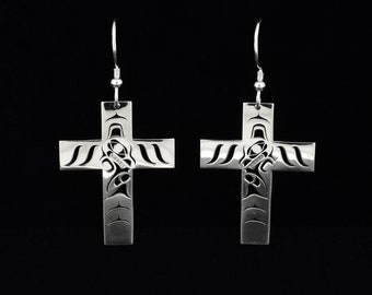 May include: A pair of silver cross earrings with intricate Native American-inspired designs. The crosses have a detailed pattern in the center and along the arms, suspended from silver ear wires against a black background.