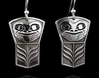May include: A pair of silver-toned earrings with a stylized Native American design. Each earring features a detailed face at the top and a geometric pattern at the bottom. The earrings hang from a simple hook.