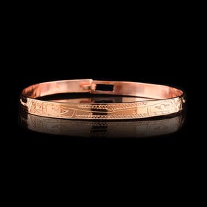 May include: A rose gold bangle bracelet with an intricate engraved design. The design features geometric patterns and stylized figures.