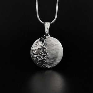 May include: A silver pendant necklace with a circular pendant. The pendant has a detailed design of a face or mask on one side and a textured, striped pattern on the other. The necklace chain is a thin, silver snake chain.