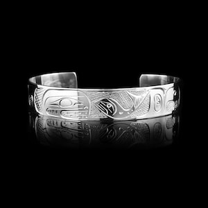 Op de afbeelding: Een zilveren manchetarmband met ingewikkelde gegraveerde ontwerpen. De armband heeft gedetailleerde afbeeldingen van gestileerde dieren en geometrische patronen. Het gepolijste metaal reflecteert de donkere achtergrond en benadrukt het gegraveerde kunstwerk.