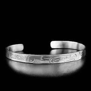 Puede incluir: Una pulsera tipo brazalete de plata con un patrón grabado de figuras animales estilizadas con líneas intrincadas y texturas entrecruzadas. La pulsera tiene un diseño abierto y un acabado metálico pulido con tallas detalladas.
