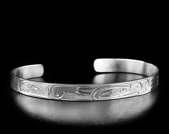 May include: A silver cuff bracelet featuring an engraved pattern of stylized animal figures with intricate lines and cross-hatched textures. The bracelet has an open back design and a polished metallic finish, showcasing detailed artistic carvings.