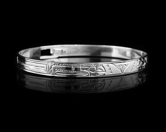 May include: A silver bangle bracelet with intricate engraved designs. The bracelet features geometric and stylized patterns along its surface. The polished metal reflects light, creating highlights and shadows against the black background. A classic piece of jewelry.