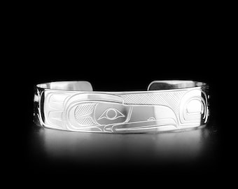 May include: A polished silver cuff bracelet featuring an intricate engraved design of a bird with stylized wings and geometric patterns. The metal surface is reflective, showcasing deep, precise lines that form the detailed avian motif.