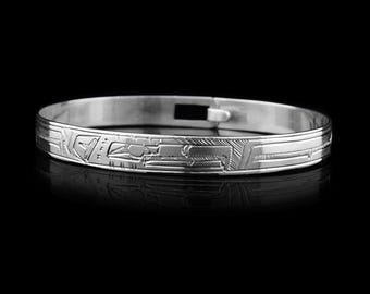 May include: A silver-colored bangle bracelet with intricate engraved designs. The bracelet features a clasp closure and detailed patterns across its surface. The design appears to be of Native American origin, with geometric and symbolic elements.
