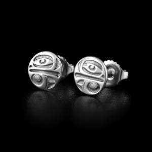 May include: A pair of silver stud earrings. Each earring has a circular design with an engraved, stylized face. The design includes eye and mouth shapes, with a tribal art aesthetic. The earrings are set against a black background.
