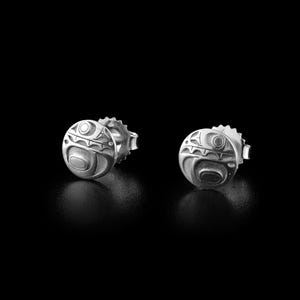 May include: A pair of silver stud earrings with a detailed, circular design. Each earring features a stylized, engraved pattern with curved lines and shapes, set against a black background. The earrings have a post and backing for secure wear.