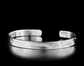 May include: A silver bracelet with an intricate design of stylized flowers and birds. The bracelet is made of polished metal and has a smooth, rounded surface.