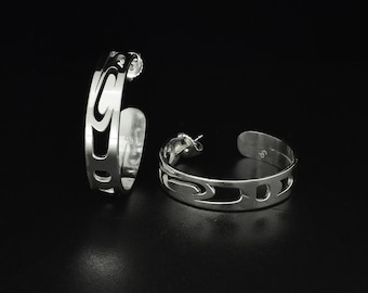 May include: A pair of silver hoop earrings with a cut-out design. The earrings have a curved shape and a post closure. The design features a series of curved and rectangular cut-outs. The earrings are displayed against a black background.