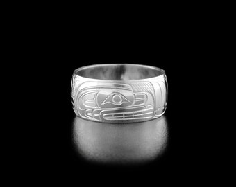 May include: A silver ring with a detailed carving of a stylized animal face. The ring is wide and has a smooth, polished finish.