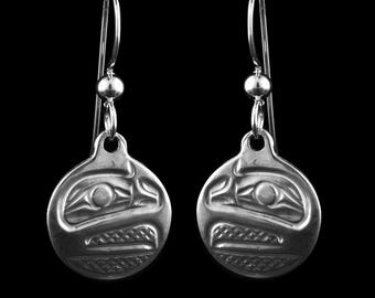 May include: A pair of silver-toned earrings with a circular pendant featuring a detailed, stylized bird design. The earrings have a hook closure and a small bead detail. The background is solid black.