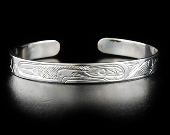 May include: A silver bracelet with a detailed engraved design of a bird, possibly a raven or eagle, in a traditional Northwest Coast style.