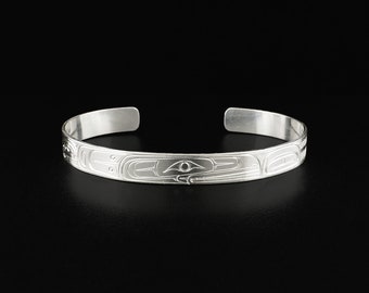 May include: A silver bracelet with a detailed design of a stylized animal face. The design is in a Northwest Coast style.
