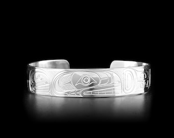 May include: A polished silver cuff bracelet featuring intricate engraved line work that depicts stylized animal forms. The metal surface is reflective and smooth, showcasing detailed patterns that wrap around the exterior of the open-ended band.