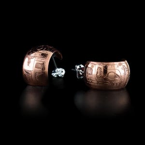 May include: A pair of copper-coloured half-hoop earrings with detailed etched designs. The earrings have a curved shape and are set against a black background. The etched patterns appear to be geometric and may represent a cultural motif.