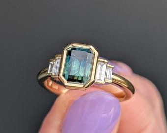 May include: A gold ring featuring a large, emerald-cut teal gemstone, flanked by smaller, rectangular white gemstones. The ring has a classic design with a polished gold band and a bezel setting for the central stone.
