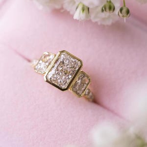 May include: A three-stone ring with a gold band and three rectangular, clear gemstones. The ring is set in a pink jewelry box, with white flowers in the background. The gemstones are the focal point of the ring.
