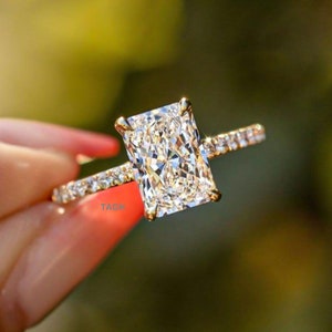 May include: A gold engagement ring with a large, rectangular, clear diamond. The diamond is set in a prong setting and the band is a thin, pave-set band.