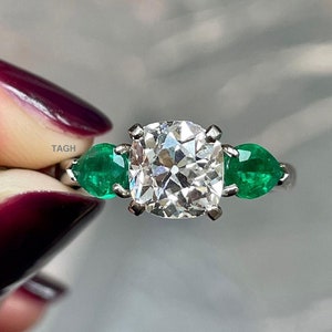 May include: A silver ring with a large, square-cut diamond and two emerald green stones on either side.
