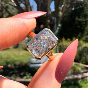 May include: A gold ring with a large, rectangular, clear gemstone. The gemstone has a multifaceted appearance, reflecting light. The ring is held up by a person's fingers, with a blurred background of greenery.