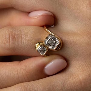 May include: A gold ring featuring two square-cut, diamond-like stones. The ring's design includes a curved band that connects the two stones. The diamonds are set in a square shape with a smaller square inside.