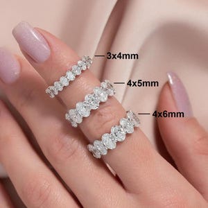 May include: Three diamond eternity rings, each with a different oval-cut stone size: 3x4mm, 4x5mm, and 4x6mm. The rings are displayed on a person's finger against a soft pink background. The rings are made of a shiny, silver-colored metal.